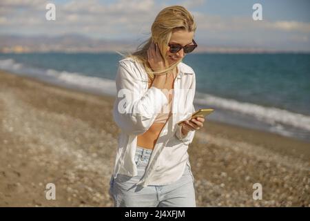 Content donna che riceve un messaggio, utilizzando lo smartphone mentre passeggi lungo la spiaggia Foto Stock