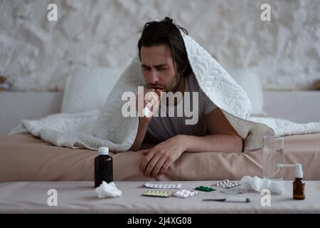 Triste uomo millenario europeo stanco con stoppia si trova sotto le coperture, tosse e trattati in camera da letto interno con pillole Foto Stock