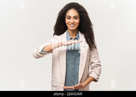 Sorridente giovane afroamericana con capelli afro indossando casual elegante su sfondo bianco tenendo vuoto con le mani, misurare il simbolo, sorridendo e guardando la macchina fotografica, concetto di pubblicità Foto Stock