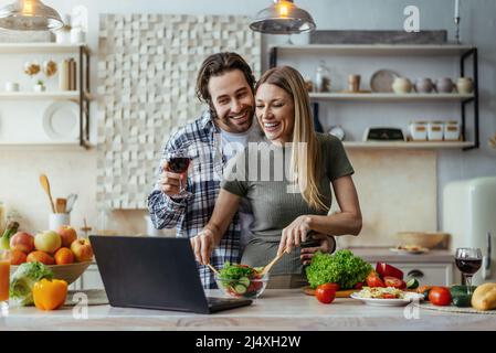 Soddisfatto marito caucasico millenario con stubble e moglie cucinare insieme, bere vino e parlare con il computer portatile Foto Stock