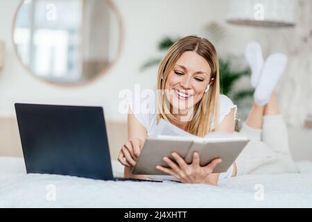 Sorridente giovane donna bionda europea in cuffie wireless lettura libro o notebook, sdraiato a letto Foto Stock