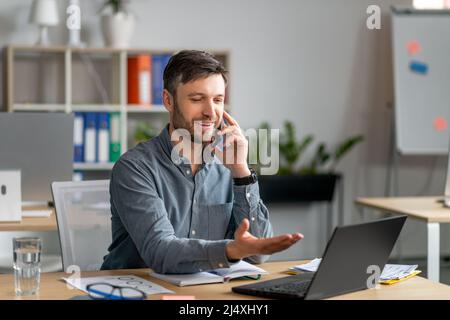 Uomo d'affari felice maturo che parla sul cellulare, discute di domande di lavoro, guarda il laptop, seduto sul posto di lavoro Foto Stock
