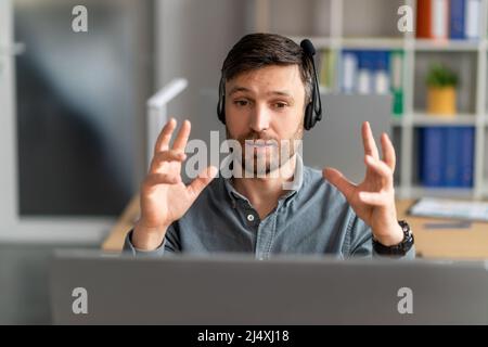 Ritratto di un manager maschile concentrato in cuffia con chiamata Web su pc seduto alla scrivania sul posto di lavoro, spiegando la strategia Foto Stock