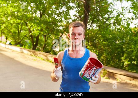 Il ragazzo dipinge il muro con un pennello in colori luminosi, tiene una lattina di vernice in mano, sulla strada Foto Stock