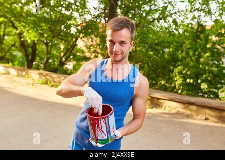 Il ragazzo dipinge il muro con un pennello in colori luminosi, tiene una lattina di vernice in mano, sulla strada Foto Stock