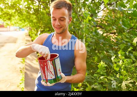 Il ragazzo dipinge il muro con un pennello in colori luminosi, tiene una lattina di vernice in mano, sulla strada Foto Stock