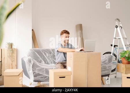 Un giovane seduto sul divano nella sua nuova casa usando il suo laptop. Foto Stock