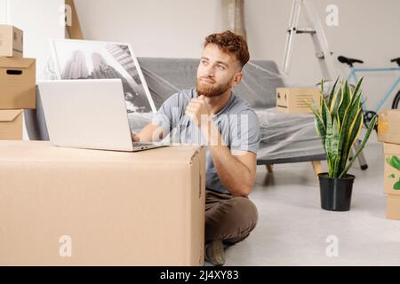 Un giovane seduto sul pavimento della sua nuova casa utilizzando il suo laptop. Foto Stock