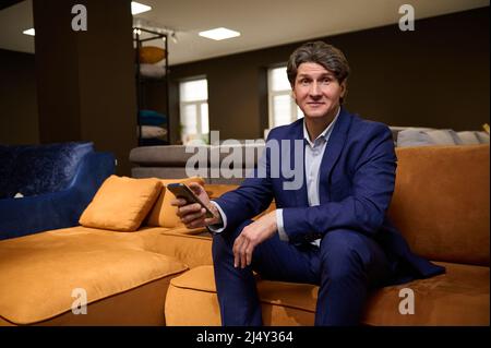 Ritratto di un uomo caucasico maturo che fa shopping o lavora in un magazzino di mobili imbottiti, seduto su un divano con smartphone nelle sue mani e guardando Foto Stock