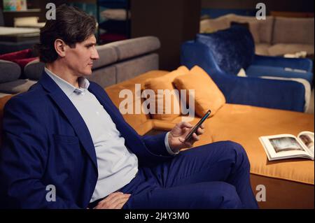 Sicuro uomo caucasico maturo che tiene il telefono cellulare, leggere messaggi, scorrere e navigare su siti internet mentre si siede su un comodo divano wi Foto Stock