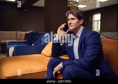 Ritratto di un bel uomo europeo maturo in tuta aziendale, cliente acquirente parlare sul telefono cellulare mentre si visita un negozio di mobili e testin Foto Stock