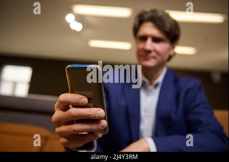 Primo piano delle mani di un uomo d'affari maturo sfocato, responsabile delle vendite che tiene un telefono cellulare, digitando messaggi di testo, navigando su siti web, navigando, swip Foto Stock