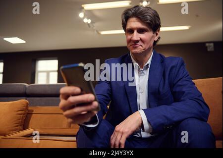 Ritratto di un uomo europeo maturo che fa shopping o lavora in un magazzino di mobili imbottiti, seduto su un divano con smartphone nelle sue mani e guardando un Foto Stock
