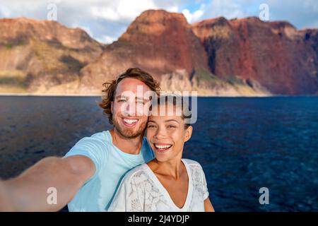 Happy coppia turisti in crociera al tramonto a Na Pali Coast Kauai, Hawaii scattando foto selfie con app per telefono cellulare. Sorridente donna asiatica e uomo caucasico Foto Stock