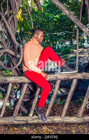 Pensando all'esterno. Vestirsi in maglione arancione chiaro con colletto alto, pantaloni rossi, scarpe con motivi geometrici, un giovane ragazzo nero è seduto sul recinto, piegando l Foto Stock