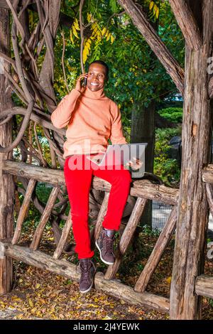 Uomo che lavora all'esterno. Vestendo in maglione arancione chiaro con colletto alto, pantaloni rossi, scarpe con motivi geometrici, un giovane ragazzo nero è seduto sul recinto, smilin Foto Stock