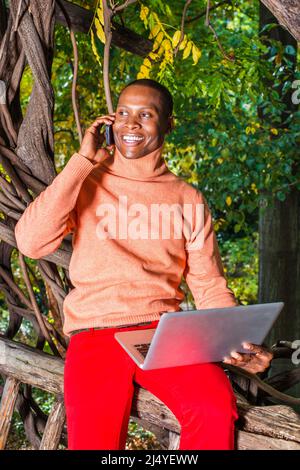 Uomo che lavora all'esterno. Vestendo in maglione arancione chiaro con colletto alto, pantaloni rossi, un giovane ragazzo nero è seduto su area di legno, sorridente, facendo telefono c Foto Stock