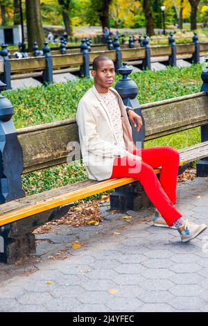 Uomo che ti aspetta. Vestirsi in blazer crema, maglione senza collari, pantaloni rossi, sneakers, un giovane ragazzo nero è seduto sulla panchina del parco, lookin Foto Stock