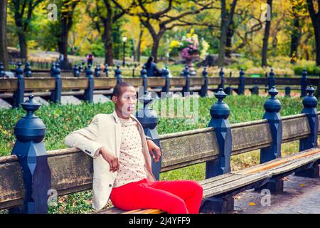 Uomo che ti aspetta. Vestirsi in blazer crema, maglione senza collari, pantaloni rossi, un giovane ragazzo nero è seduto sulla panchina del parco, braccia stretching Foto Stock
