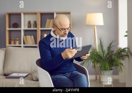 Uomo anziano serio in occhiali seduti in sedia e utilizzando il suo moderno tablet digitale Foto Stock