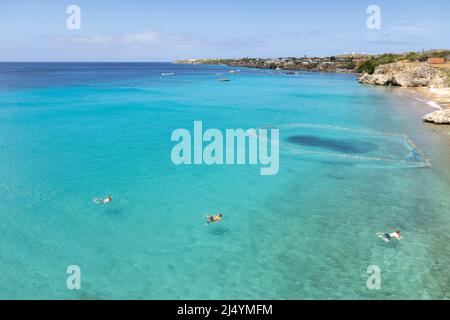 Tre ragazzi che tornano alla spiaggia dopo aver saltato da una scogliera nel mare dei Caraibi con una rete da pesca e barche sullo sfondo; Curacao Foto Stock