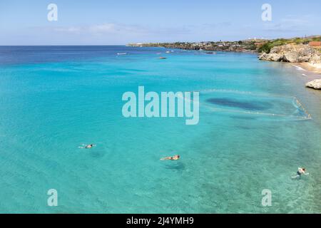 Tre ragazzi che tornano alla spiaggia dopo aver saltato da una scogliera nel mare dei Caraibi con una rete da pesca e barche sullo sfondo; Curacao Foto Stock