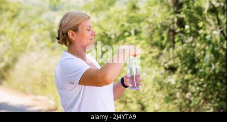 Vista laterale foto di una donna bionda all'aperto nella foresta con t-shirt bianca e smart watch cercando di aprire la bottiglia d'acqua. Foto Stock