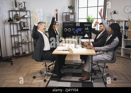 Sorridendo diversi uomini d'affari allegri e multirazziali si siedono alla scrivania dell'ufficio, celebrando il progetto di successo, gesturando, ridendo, stringendo le mani e condividendo insieme le idee di affari. Foto Stock