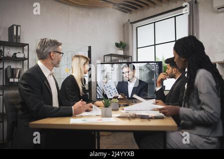 Sicuro uomo caucasico di mezza età dice il concetto di programma di affari ai suoi colleghi multinazionali su un grafico durante una videoconferenza in un m Foto Stock