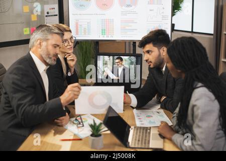 Concetto di riunione elettronica. Diversi gruppi di uomini d'affari multirazziali nella sala conferenze ascoltano le istruzioni dei loro colleghi d'affari durante una videoconferenza da un computer portatile. Foto Stock