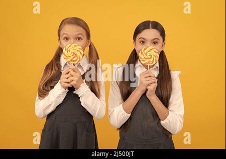 I bambini affamati della scuola tengono i lollipops colorati di swirl sui bastoni sfondo giallo, cibo Foto Stock