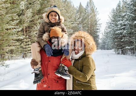 Famiglia allegra di tre in winterwear caldo che passa la giornata nella foresta invernale o parco tra firtrees e pini coperti di neve Foto Stock