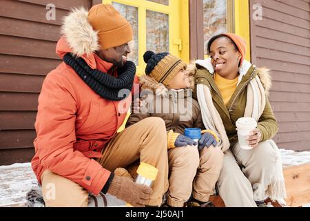 Allegra donna nera in winterwear guardando il suo figlio carino durante la conversazione mentre si siede sul portico del cottage vicino al giovane uomo e avendo il tè Foto Stock