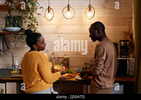 Giovane coppia contemporanea afroamericana in pulllovers preparare la colazione insieme nella cucina della loro casa di campagna Foto Stock
