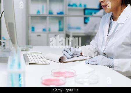 Mani di giovane scienziata femminile in whitecoat facendo appunti in documenti clinici mentre si siede sul posto di lavoro in laboratorio Foto Stock