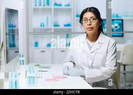 Giovane virologo femminile serio o microbiologo in whitecoat guardando la macchina fotografica mentre si siede sul posto di lavoro con provette e piastre Petri Foto Stock