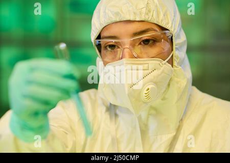 Scienziata femminile in occhiali protettivi, respiratore e tuta a a rischio biologico che guarda il campione chimico nella provetta in mano con i guanti Foto Stock