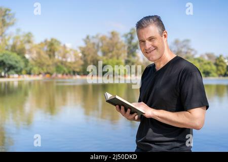 Uomo maturo che legge su un lago, godendo del tempo libero in una giornata di sole Foto Stock