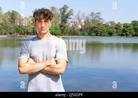 Ritratto di giovane biondo fiducioso con braccia incrociate guardando la macchina fotografica, sfondo lago estivo. Spazio di copia Foto Stock