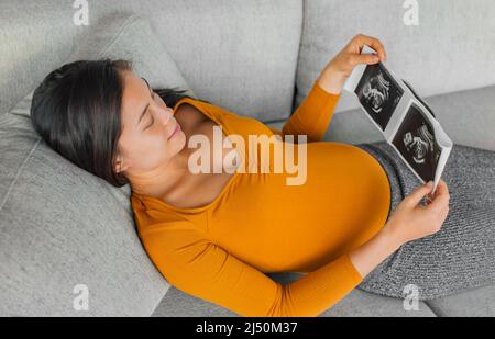 Scansione ecografica del bambino. Donna asiatica incinta che tiene l'immagine dell'ospedale risultato da controllo di salute con il ventre piccolo felice di prima gravidanza Foto Stock