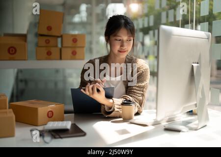 Giovane donna piccola impresa proprietario online shopping a casa. Elenco ordini da clienti con cellulari preparazione pacchetto prodotto in background. PMI Foto Stock