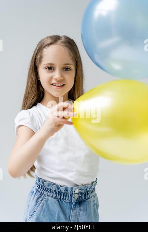 ragazza felice guardando la macchina fotografica vicino blu sfocato e palloncini gialli isolato sul grigio Foto Stock
