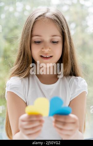 sorridente ragazza patriottica con capelli lunghi che tengono blurred blu e giallo cuori di carta Foto Stock