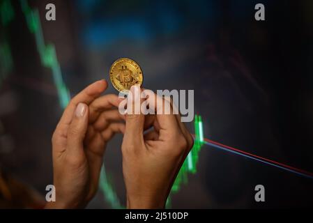 Moneta di criptovaluta Bitcoin nelle mani femminili contro i grafici, tassi su sfondo sfocato. Foto Stock