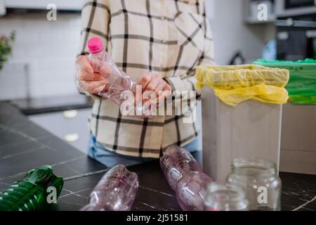 Sezione centrale della donna gettando una bottiglia di plastica vuota in un contenitore di riciclaggio in cucina. Foto Stock