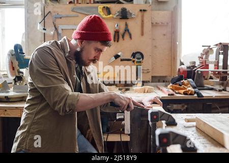 Vista laterale del carpentiere caucasico con cappuccio in maglia rossa che taglia la mortasa in legno con scalpello Foto Stock