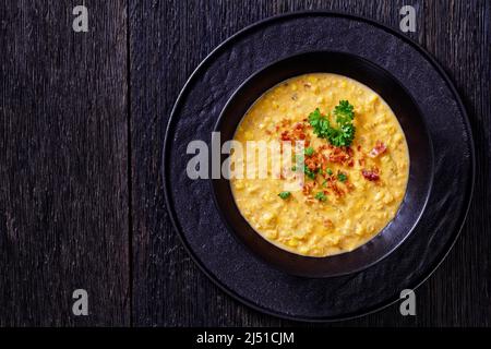 zuppa di mais con pancetta fritta croccante sbriciolata in una ciotola nera su un tavolo di legno scuro, vista orizzontale dall'alto, posa piatta, spazio libero, primo piano Foto Stock