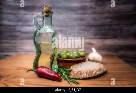Piano tavolo ancora vita di olio ovlive, olive verdi, peperoncino rosso, rametto di rosmarino, aglio e pane in un ambiente rustico. Foto Stock