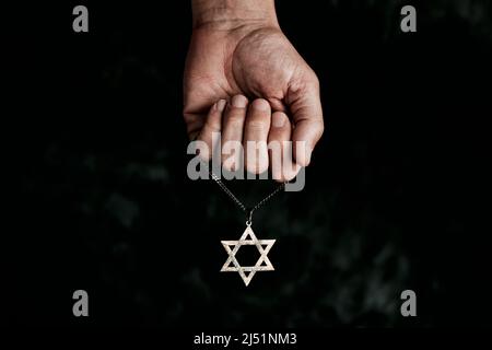 primo piano di un vecchio e arrugginito pendente a forma di stella di davide in mano di un uomo su sfondo nero chiazzato Foto Stock