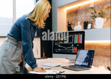 Donna bionda caucasica matura e di successo, un commerciante di azioni, un investitore criptato, si trova vicino al desktop, analizza le carte su un computer e schermo di un laptop, confronta gli indicatori, valuta i rischi Foto Stock